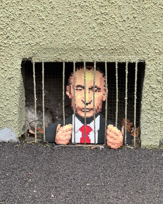 Street art by Swedish artist Johan Karlgren, known as Pappas Pärlor, featuring a pixelated depiction of Vladimir Putin behind bars. The artwork is integrated into a real street vent in Motala, Sweden, using Karlgren's signature old-school video game-inspired pixel art style. The piece shows Putin's face and hands gripping the bars, symbolizing imprisonment and accountability. This interactive and humorous work adds a powerful voice to the global protest against the war in Ukraine, blending playful aesthetics with serious political commentary