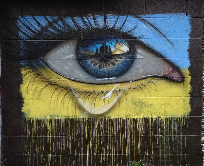 Street art mural by My Dog Sighs featuring a detailed, teary eye against the backdrop of the Ukrainian flag. The vibrant blue iris reflects the silhouette of a Ukrainian cityscape at sunset, symbolizing resilience and hope. A single tear rolls down, and yellow paint drips from the flag-inspired background, representing sorrow for the hardships faced by Ukraine. This mural is a poignant tribute to the strength and spirit of the Ukrainian people during times of conflict.