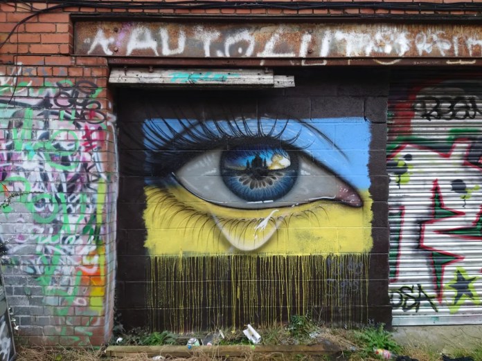 Street art mural by My Dog Sighs, painted on a weathered brick wall amidst graffiti in an urban environment. The artwork depicts a detailed, teary eye with a vibrant blue iris reflecting the silhouette of a Ukrainian cityscape at sunset. The yellow background beneath the eye, representing the Ukrainian flag, features dripping paint, symbolizing sorrow and resilience. Surrounding the mural are layers of colorful graffiti tags and a rusted, dilapidated shutter, enhancing the raw and gritty atmosphere of the setting. This piece is a tribute to the strength and endurance of the Ukrainian people during times of conflict.