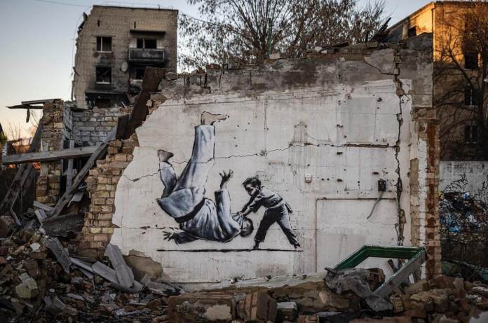 Street art mural by Banksy on the wall of a war-torn building in Ukraine, depicting a young boy in a judo match throwing a man to the ground, widely interpreted as a representation of Vladimir Putin. The mural is painted on a heavily damaged structure surrounded by rubble and debris, with a destroyed apartment building visible in the background. The scene symbolizes the defiance and strength of Ukraine against oppression, using Banksy's signature monochromatic style with powerful symbolism. The setting emphasizes the devastating impact of the ongoing conflict.