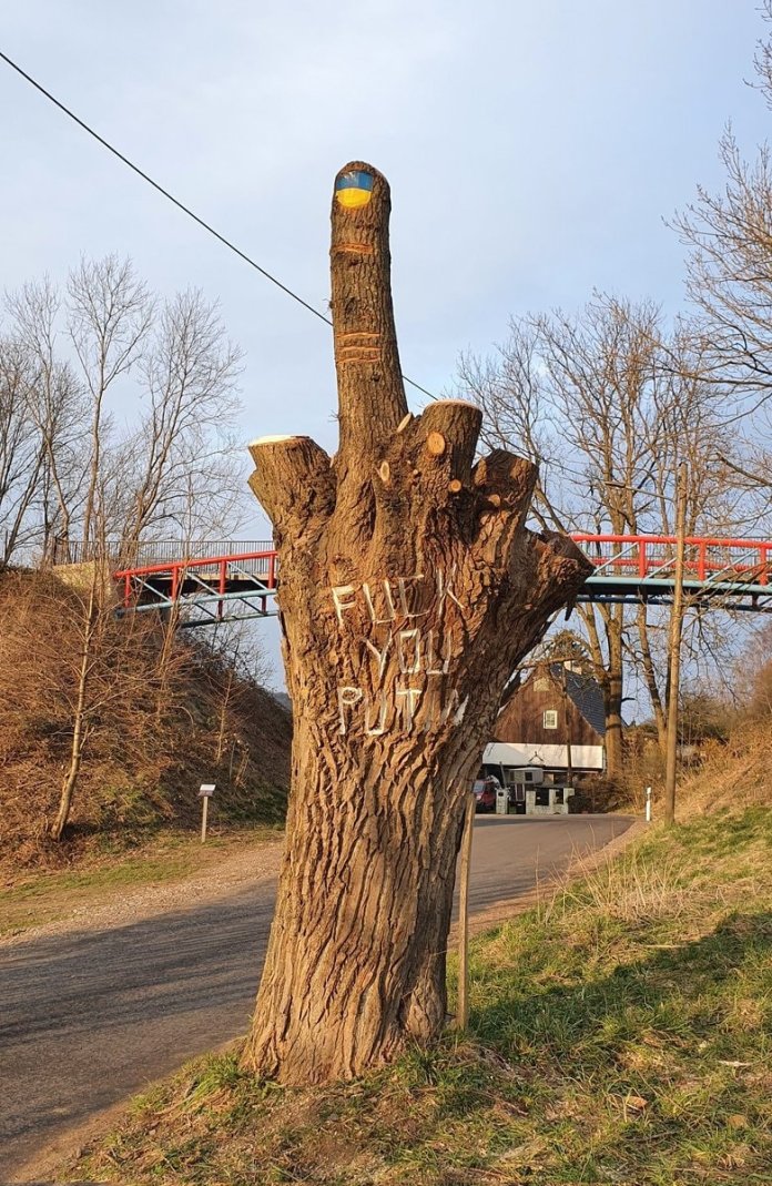 A modified tree stump creatively carved and painted to depict a hand making an obscene gesture with its middle finger raised, symbolizing defiance against Vladimir Putin. The top of the middle finger is painted in the blue and yellow colors of the Ukrainian flag, reinforcing solidarity with Ukraine. The trunk bears the engraved words 'Fuck You Putin,' further expressing a bold and unfiltered message of protest. The scene is set along a roadside with a small bridge and house in the background, capturing a rural yet politically charged act of resistance.
