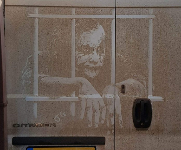 Dirty van art of the Joker in jail, leaning forward with arms hanging through dust-drawn bars, detailed in a high-contrast white-on-dirt technique by James Gibson.