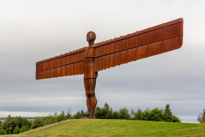 The Angel of the North sculpture in Gateshead, UK, a massive rust-colored steel figure with outstretched wings standing on a grassy hill under a cloudy sky, blending monumentally with the landscape.