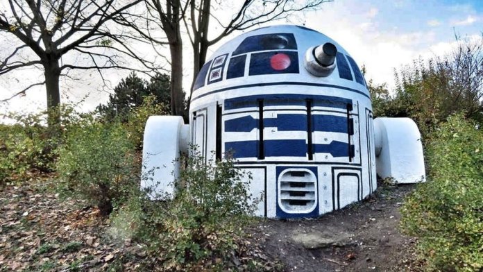 A creative piece of street art transforms a dome-shaped nuclear shelter air vent into a life-sized replica of R2-D2 from Star Wars. The structure, located in Prague, Czech Republic, is painted in the droid's signature blue, white, and silver color scheme, complete with intricate details like panels, vents, and a red eye sensor. Flanked by two white-painted side structures mimicking R2-D2's legs, the artwork blends into the surrounding landscape of trees and bushes, making it appear as if the beloved sci-fi character has landed in the forest.