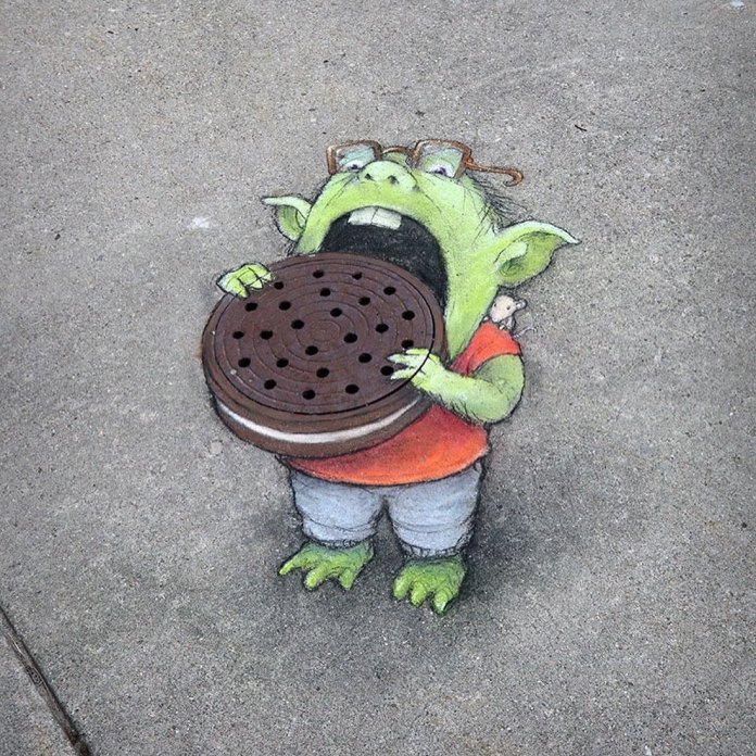 Chalk art on pavement by David Zinn showing Neil, a green goblin-like creature wearing glasses, an orange shirt, and grey shorts, holding a drain cover painted to resemble a large chocolate cookie.