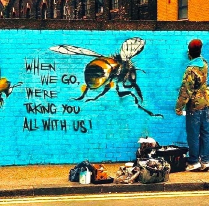 A mural by Louis Masai in London showing large bees and the words “When we go, we're taking you all with us” painted on a bright blue brick wall. The artist is seen working beside it.