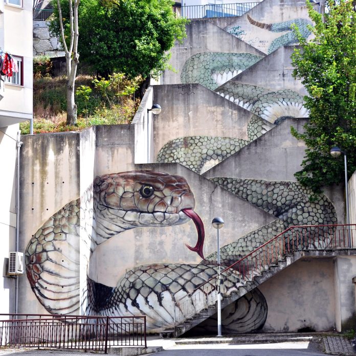 A large mural of a snake painted by SFHIR in Guarda, Portugal, uses a stairwell's vertical and diagonal lines to give the illusion of a snake coiling upward, with detailed scales and a forked tongue extending at the base.