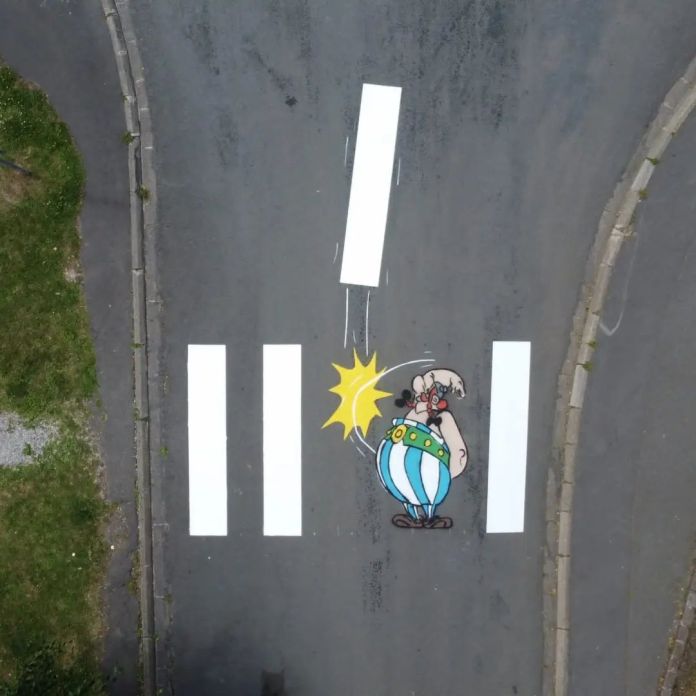 A street painting in France by Oakoak of Obelix from Asterix striking a lane line, which appears shattered, turning the crosswalk into a playful comic scene.