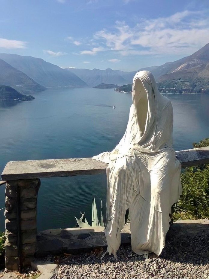 White ghost-like sculpture draped in fabric sitting on a stone bench overlooking Lake Como, created at the Castle of Vezio in Italy.