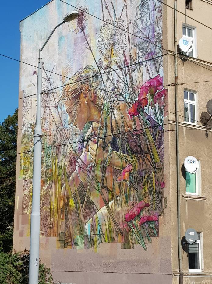 Mural by Krzysztof Bitka showing a woman kneeling in a field of tall grass, dandelions, and wildflowers. The art has soft colors and transparent, sketchy textures. Painted in Szczecin, Poland.