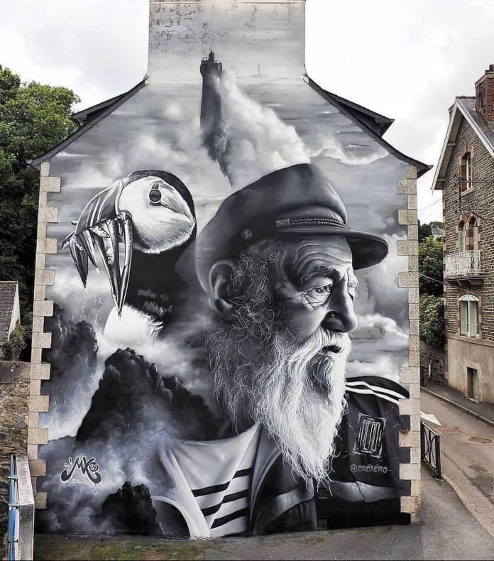 Mural by Creaero in Morlaix, France, depicting an old sailor in grayscale with a puffin, crashing waves, and a lighthouse in the distance.