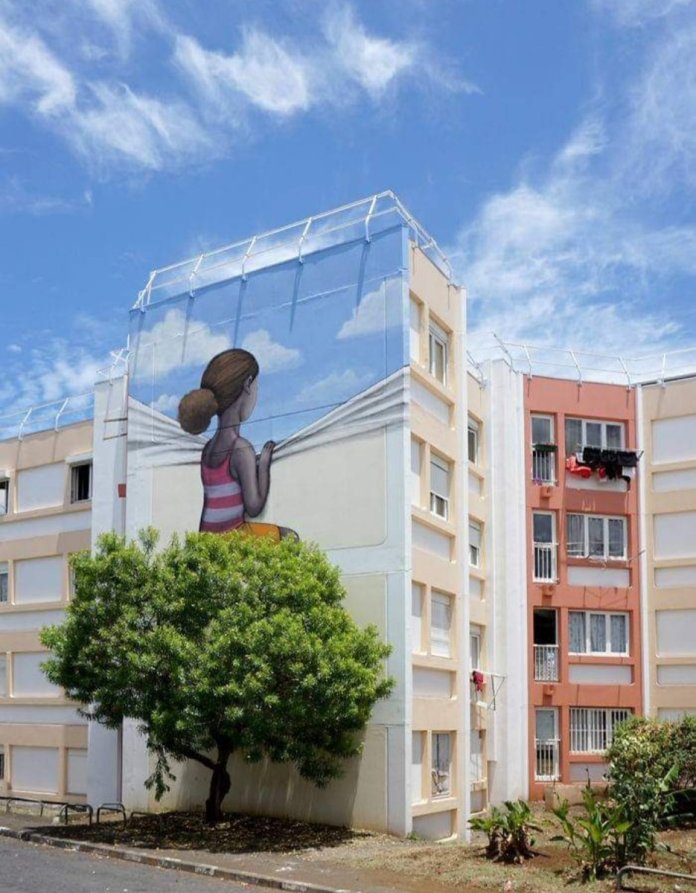 Mural by Seth Globepainter of a young girl pulling back a painted curtain of sky on a high-rise building in Le Port, Réunion Island. A real tree aligns with the artwork’s base.