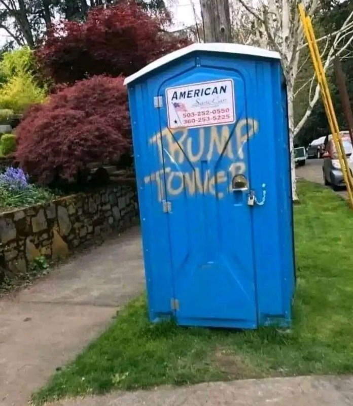 A bright blue portable toilet has “TRUMP TOWER” spray-painted in yellow across the front. It stands on a residential sidewalk near trees and grass.