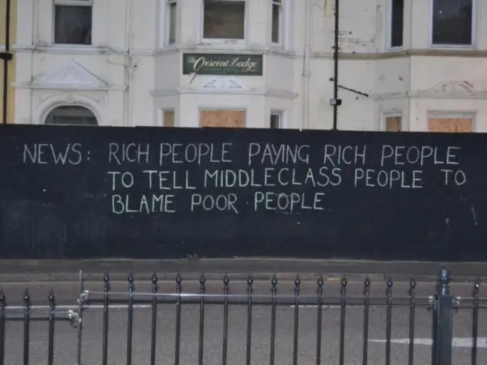 Graffiti on a black construction wall in front of a white building reads: “NEWS: Rich people paying rich people to tell middleclass people to blame poor people.” All text is in uppercase.