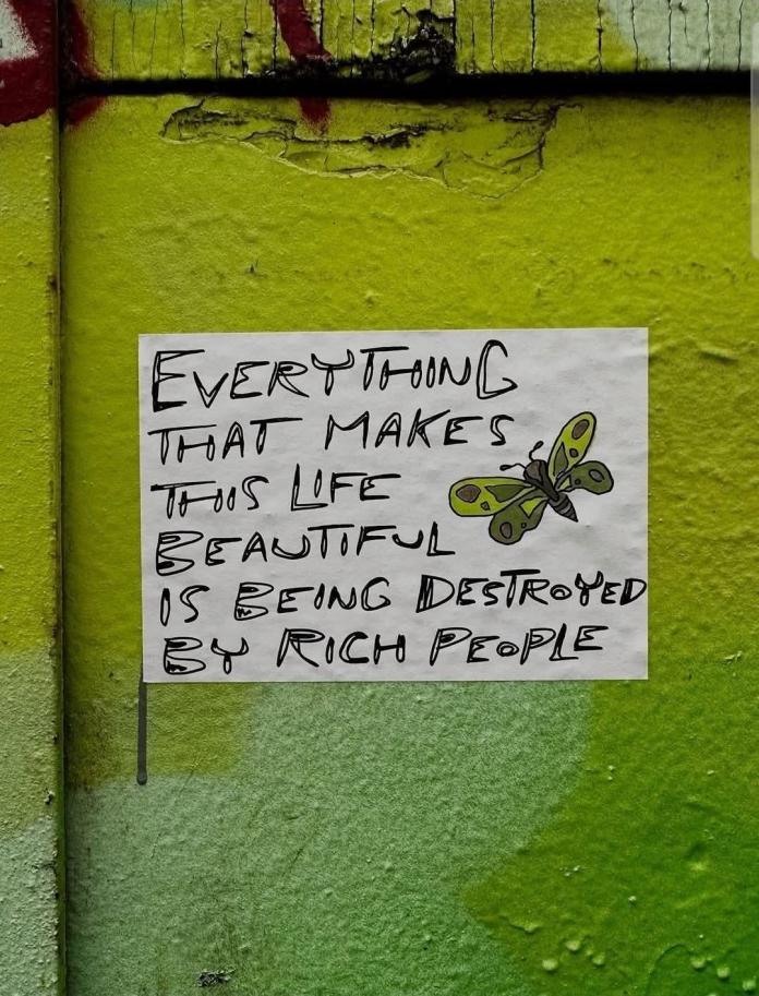 A handwritten poster on a green wall reads: “Everything that makes this life beautiful is being destroyed by rich people.” A small butterfly drawing is added on the right side of the paper.
