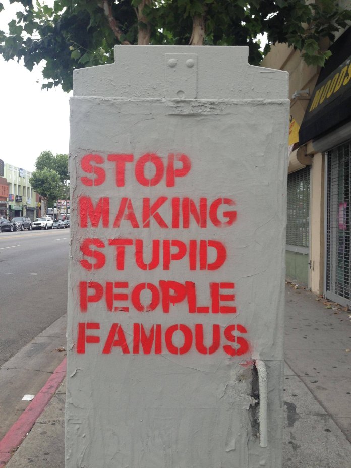 Red stencil text on a gray utility box in Los Angeles reads "STOP MAKING STUPID PEOPLE FAMOUS" in capital letters. A street scene with cars and storefronts appears in the background. Created by Plastic Jesus.
