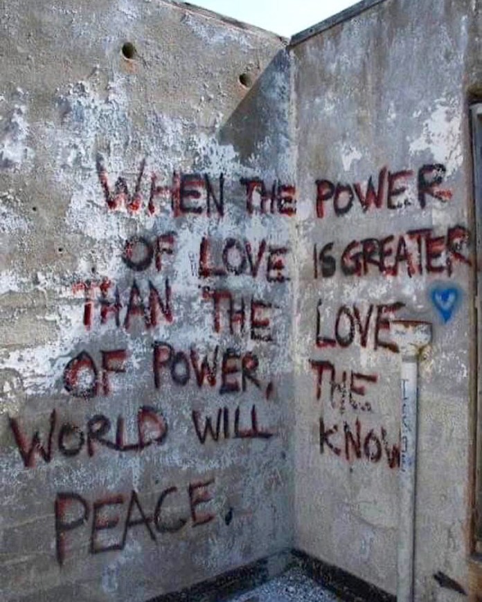 Graffiti on a weathered concrete wall reads “WHEN THE POWER OF LOVE IS GREATER THAN THE LOVE OF POWER, THE WORLD WILL KNOW PEACE” in uneven red and black letters, referencing a quote often attributed to Jimi Hendrix.