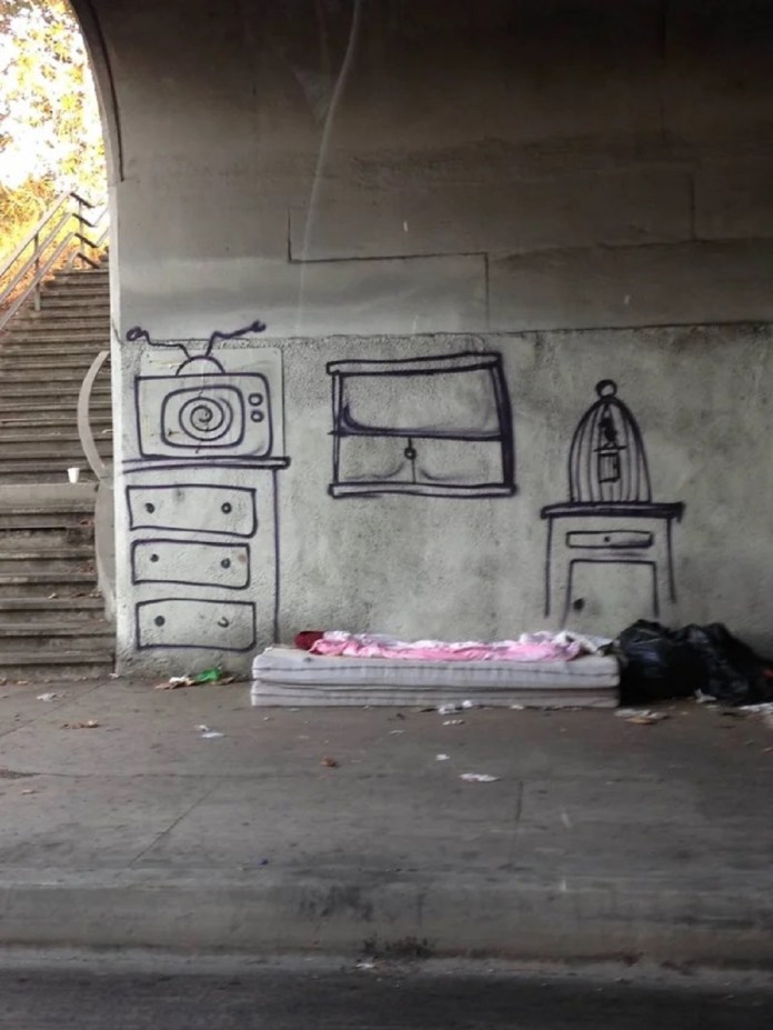 A mattress lies under a highway bridge in Los Angeles. On the wall behind it, graffiti outlines a living room scene with a window, TV, dresser, and birdcage—creating the illusion of a furnished home. Painted by Skid Robot to highlight homelessness.
