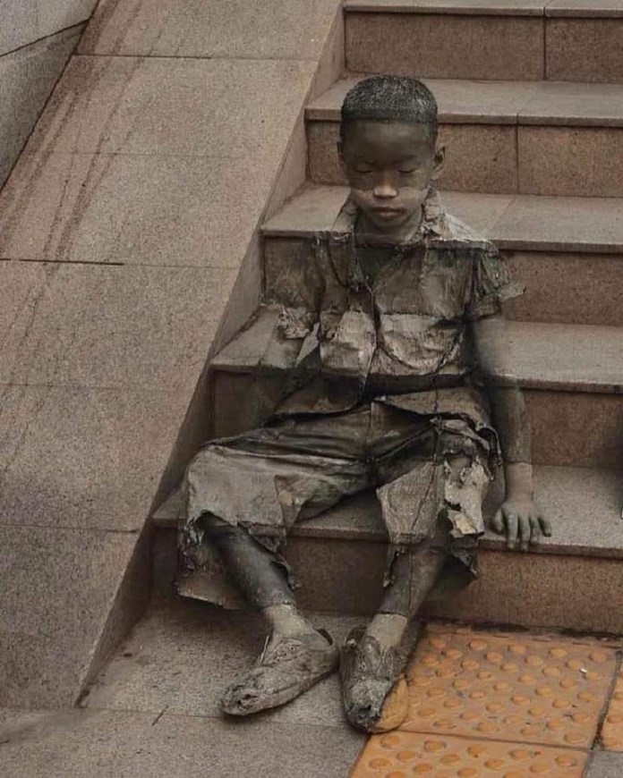 A child covered in body paint sits on concrete steps, camouflaged to blend in with the background.