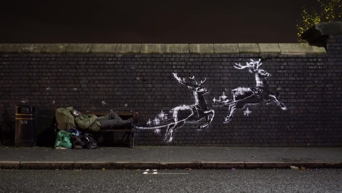 A mural by Banksy in Birmingham’s Jewellery Quarter, featuring two white reindeer painted on a brick wall. The reindeer appear to pull a public bench as if it were Santa’s sleigh. A man lies on the bench, surrounded by his belongings, highlighting the issue of homelessness. The scene is lit at night, with the reindeer depicted in Banksy’s signature stencil style.