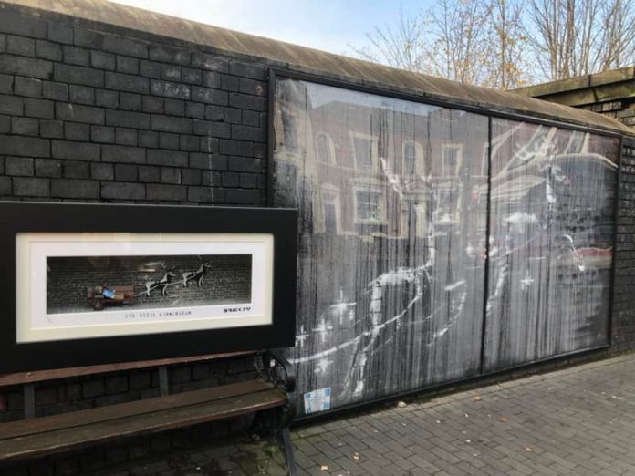 Banksy's mural in Birmingham, photographed after being covered with protective glass. The original artwork of reindeer pulling a public bench is visible behind the glass, now partially obscured by reflective surfaces. Next to the mural, a framed print of the artwork titled "God Bless Birmingham" is displayed on the bench, emphasizing the piece's ongoing impact.