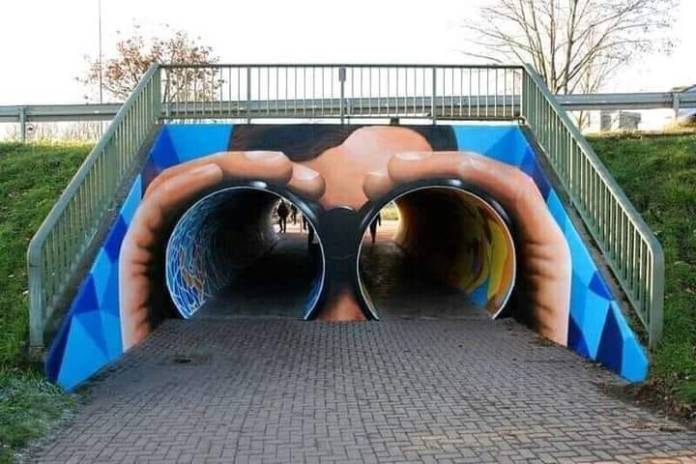 A mural painted around a pedestrian underpass transforms the twin tunnel entrances into the lenses of binoculars, with a painted person holding them up to their face. The tunnels align to complete the illusion of looking through binoculars.