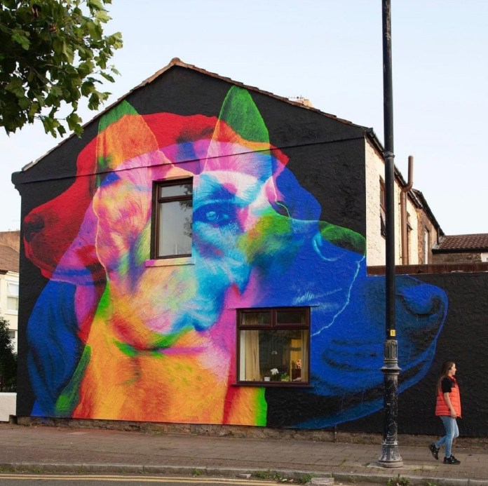 A mural by ACHES shows three overlapping dog portraits in vivid, blended colors—red, blue, green, and yellow—painted across the side of a black house in New Brighton, England. A pedestrian walks past, enhancing the scale.