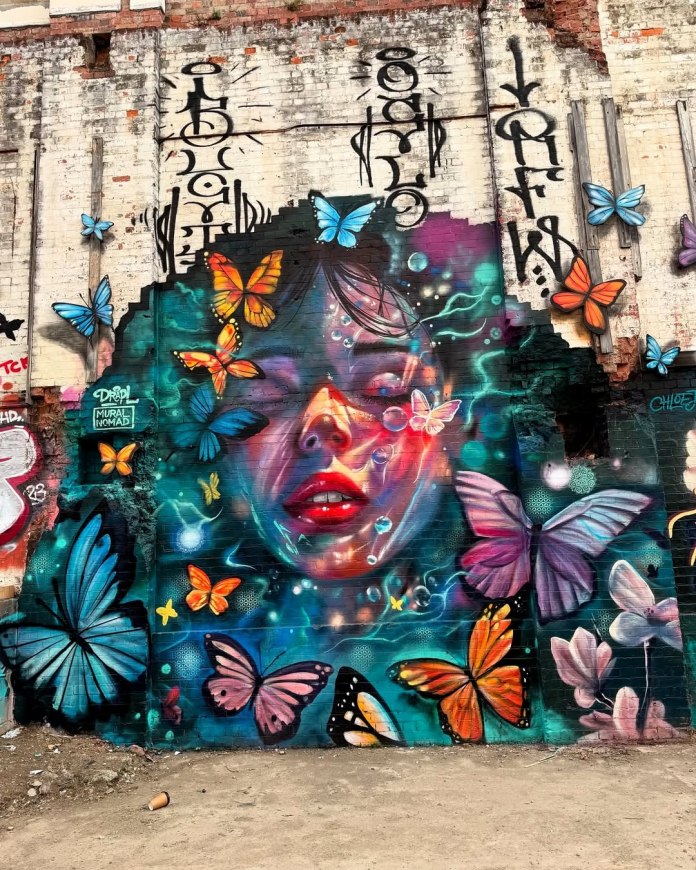 A surreal mural in Brisbane, Australia, by Drapl and Mural Nomad, portraying a woman's face blending into butterflies, painted with vibrant blues and golds against a textured background.