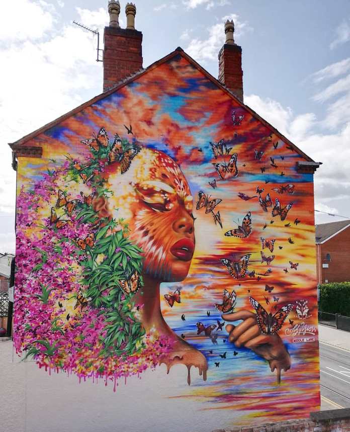 A large-scale mural by Jim Vision in Beeston, UK, showing a woman's face transforming into flowers and monarch butterflies against a sunset-colored sky.