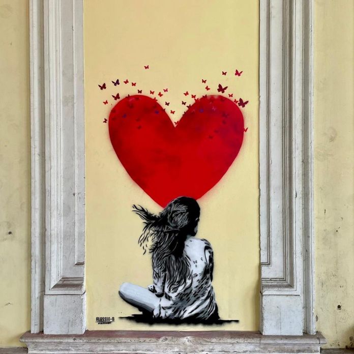 A stencil graffiti piece by Alessio-B in Montagnana, Italy, showing a seated girl looking up at a red heart breaking apart into small butterflies.