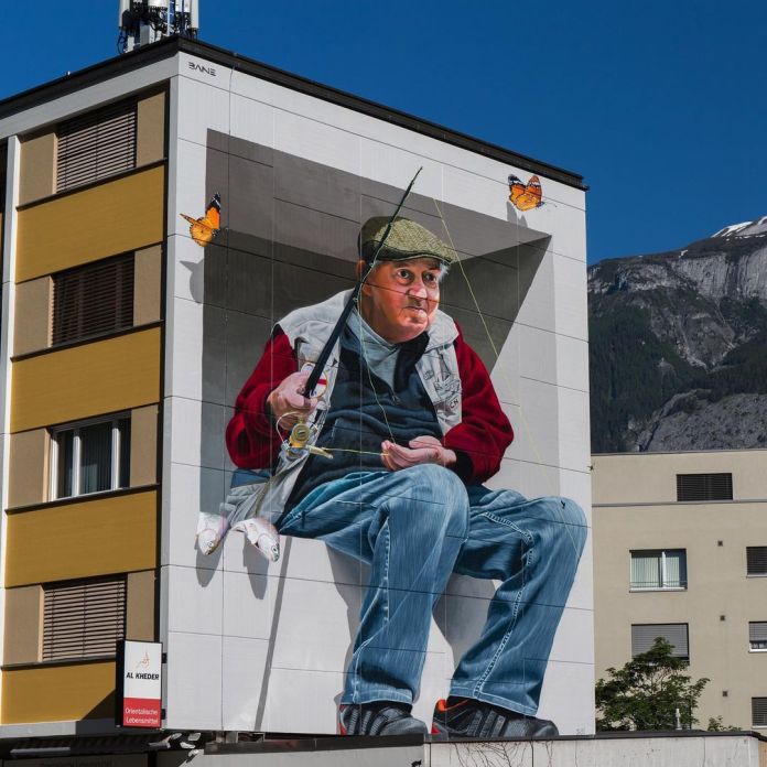 A hyperrealistic 3D mural in Chur, Switzerland, by Fabian Bane Florin, depicting an elderly fisherman sitting on a ledge with monarch butterflies fluttering around him.