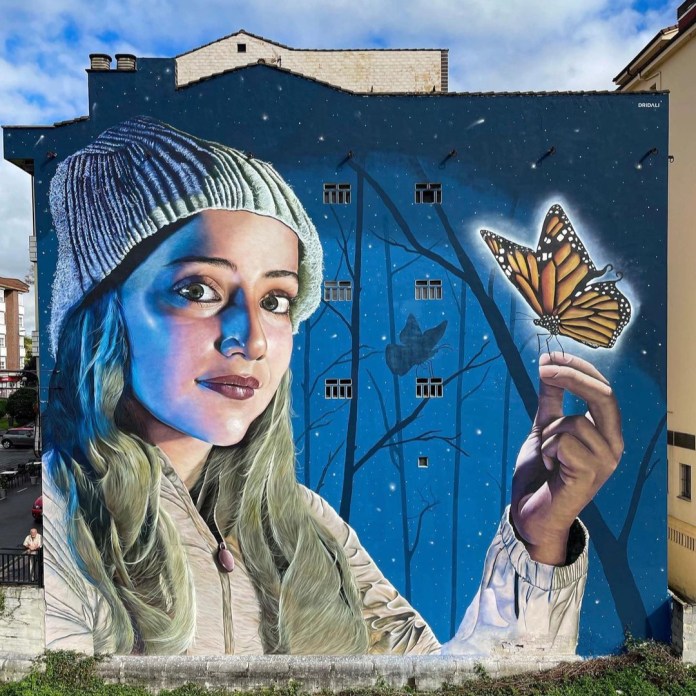 A nighttime mural in El Berrón, Spain, by Dridali, featuring a woman illuminated in blue tones, gently holding a glowing monarch butterfly.
