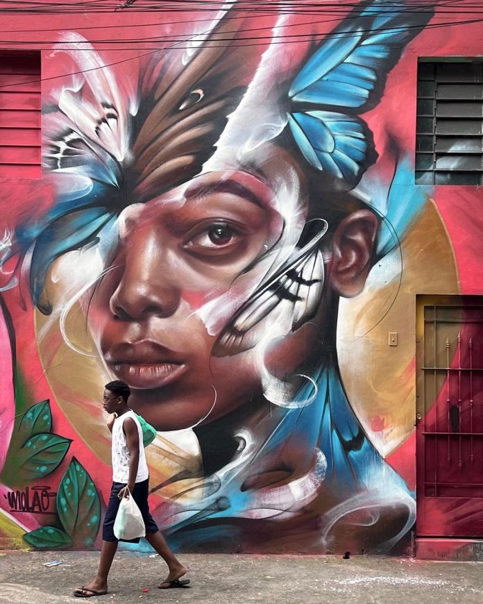 A vibrant street art mural by Filite in Taboão da Serra, Brazil, featuring a hyperrealistic portrait of a person whose face seamlessly transforms into butterflies, blending realism and surrealism in a stunning visual effect.
