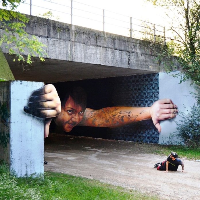 Mural under a bridge in Nerviano, Italy showing a tattooed man appearing to pull himself out of the tunnel with outstretched arms. One hand grabs the tunnel edge, while the other reaches toward an artist crawling away on the ground.