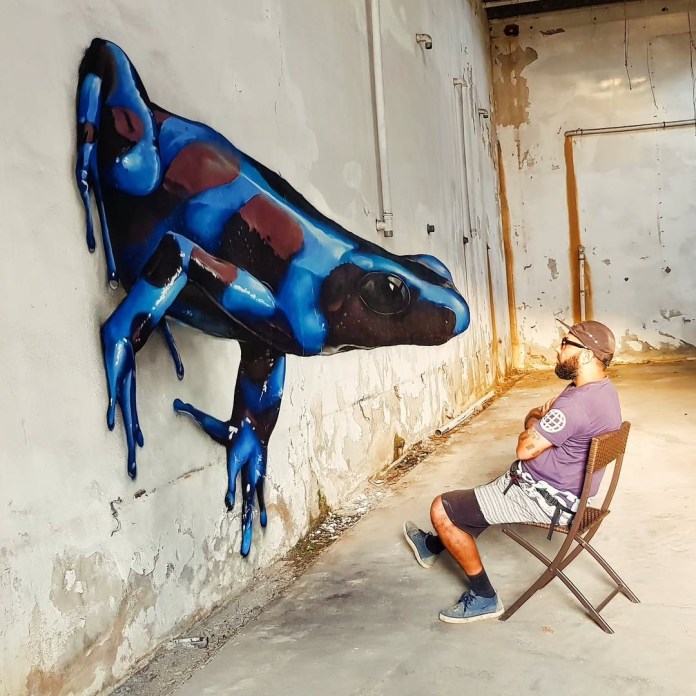 Hyperrealistic mural of a giant blue frog on a cracked interior wall in Portugal. The frog appears to emerge into the room where a man sits on a folding chair facing it.