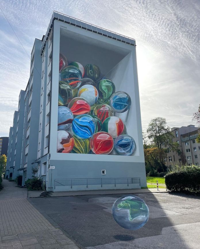 Large-scale mural in Wuppertal, Germany of a tilted box of marbles painted on a building wall. A real marble appears to roll out of the artwork onto the sidewalk in front.