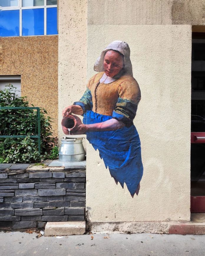 Mural of Vermeer’s milkmaid painted on a beige wall in Saint-Étienne, France. She appears to pour real liquid into a metal container placed on the sidewalk.