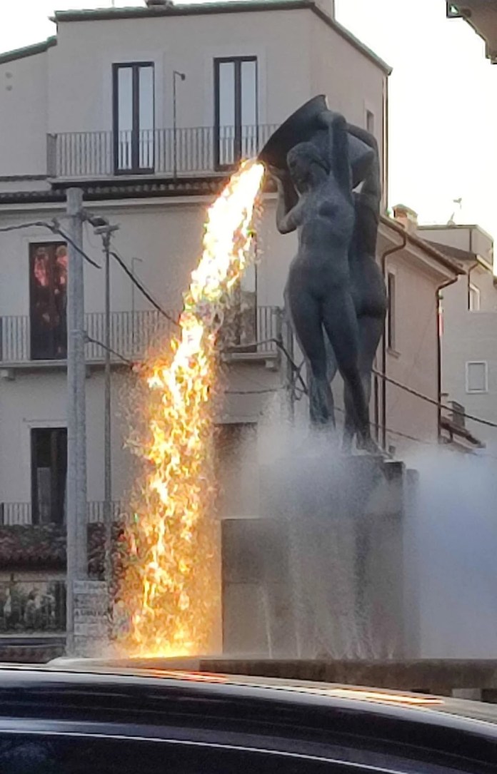 The Fontana Luminosa in L'Aquila, Italy, a sculpture by Nicola D'Antino from the 1930s, illuminated by sunlight creating an optical illusion of fiery lava pouring from the fountain. The dramatic effect highlights the stunning blend of art and nature, with surrounding buildings adding to the scene’s urban charm.