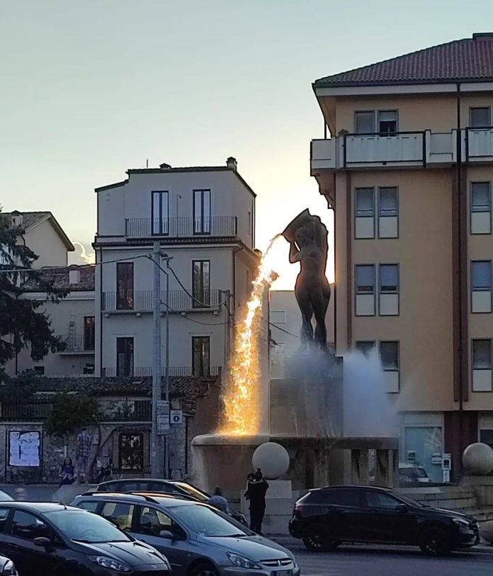 A wide-angle view of the Fontana Luminosa in L'Aquila, Italy, designed by Nicola D'Antino in the 1930s. The fountain is bathed in sunlight, creating a spectacular illusion of fiery lava streaming from the sculpture. Surrounded by buildings and evening activity, the scene captures a unique blend of urban life and artistic wonder.