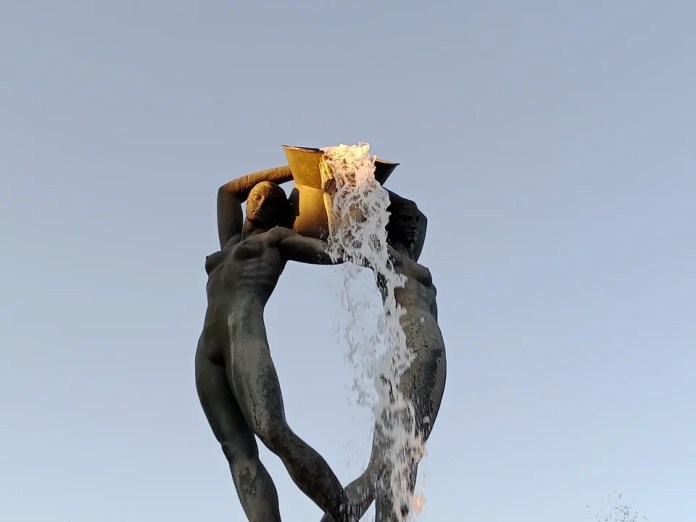 The top of the Fontana Luminosa in L'Aquila, Italy, sculpted by Nicola D'Antino in the 1930s. The sunlight illuminates the water as it cascades from the sculpture, creating the illusion of fiery lava against a clear blue sky. The details of the human figures in the sculpture are highlighted by the dramatic lighting.