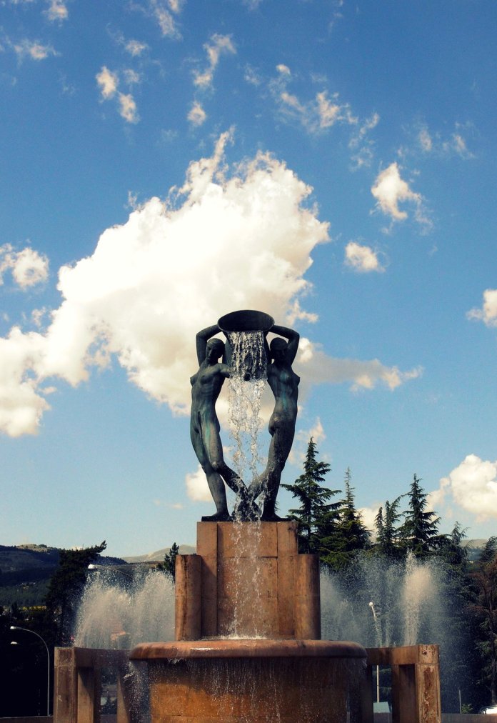 The Fontana Luminosa in L'Aquila, Italy, sculpted by Nicola D'Antino in the 1930s. The fountain is shown on a sunny day against a backdrop of a blue sky and fluffy clouds, with water pouring gracefully from the bowl held by two human figures. Trees and mountains in the distance enhance the serene and picturesque setting.