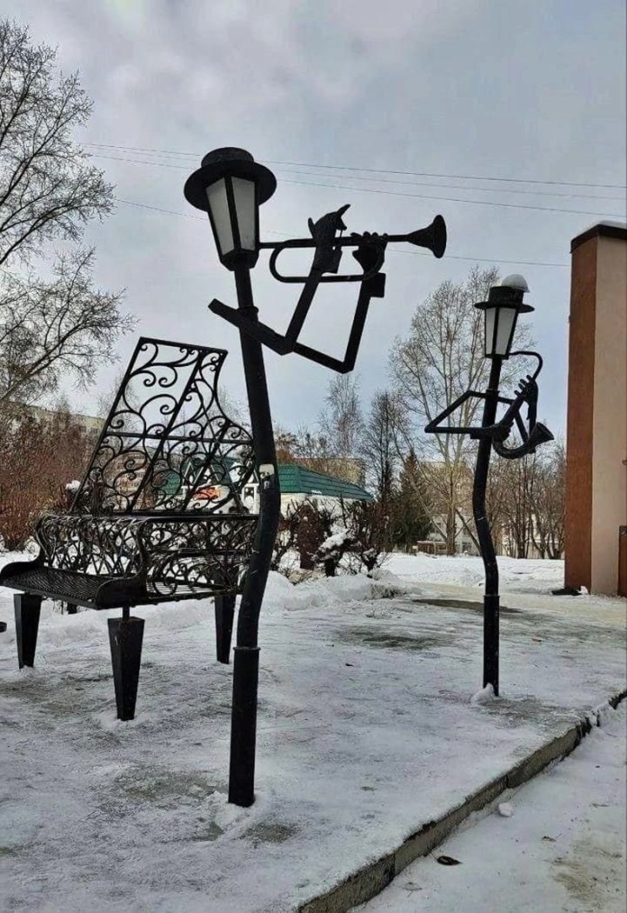 Sculptures of lamp posts shaped like jazz musicians playing trumpet and saxophone beside a piano-shaped bench in a snowy public park.