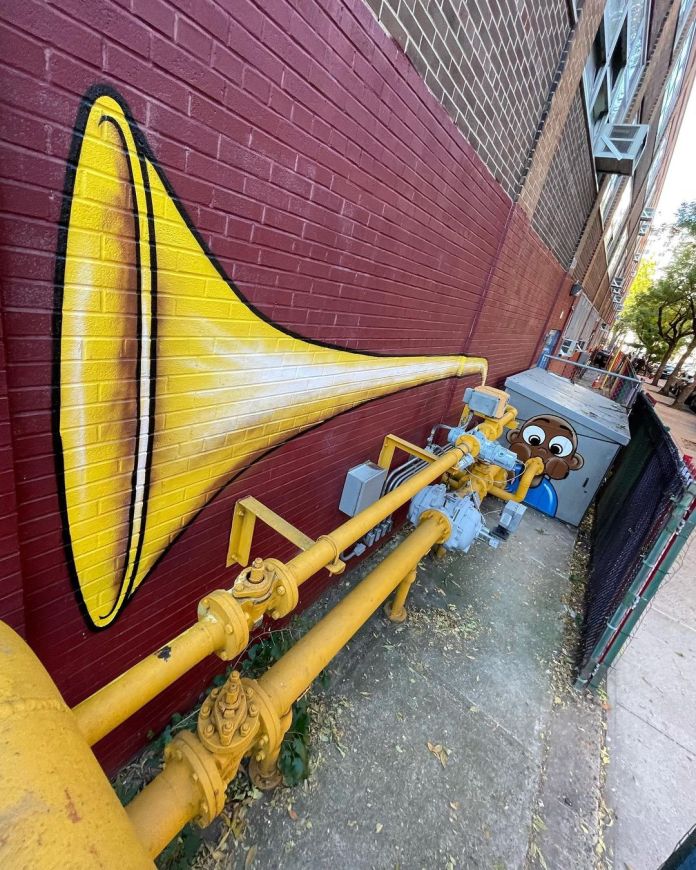 Yellow gas pipes painted as a trombone with a cartoon character blowing into it, integrated into a red brick wall outside a school in New York, USA.