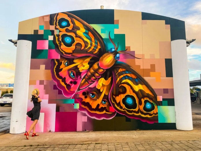 Colorful mural in Perth showing a giant butterfly with realistic wings featuring eye motifs, emerging from a pixelated background. A woman in a dress poses in front.