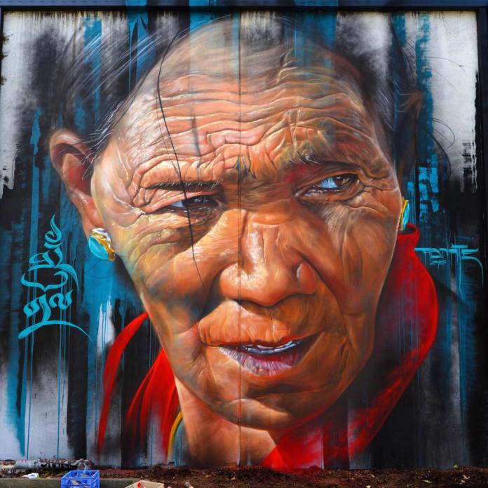 Hyperrealistic mural of an elderly Tibetan with deep wrinkles and a red scarf, painted on a vertical wall in Melbourne, Australia.