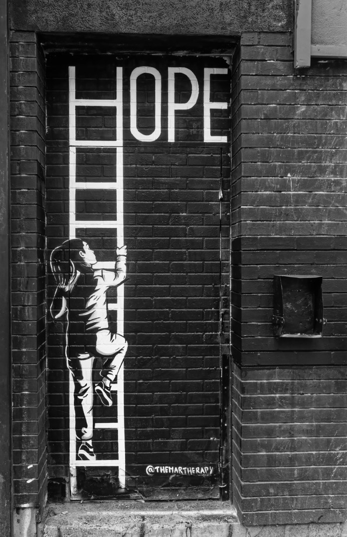 Black-and-white mural of a young girl climbing a white ladder that forms the letter H in the word “HOPE,” painted on a brick door facade in Montreal, Canada.