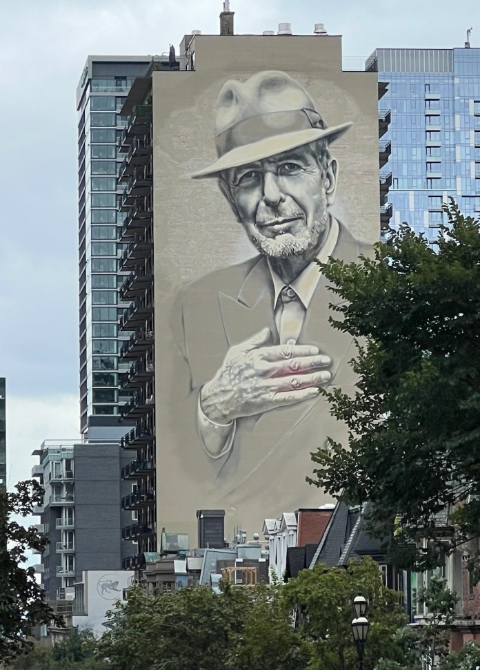 Giant mural of Leonard Cohen in a fedora, hand to heart, painted on a downtown Montreal high-rise building with modern apartments and condos surrounding the mural.