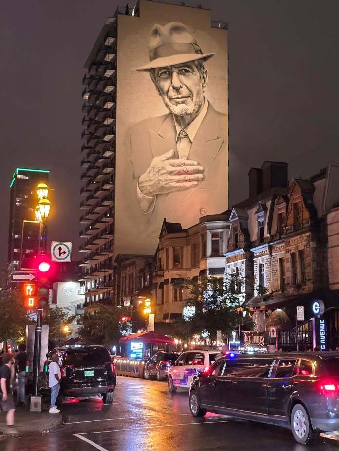 Nighttime view of the Leonard Cohen mural in Montreal, illuminated by city lights, surrounded by street life, restaurants, and traffic on a wet street corner.