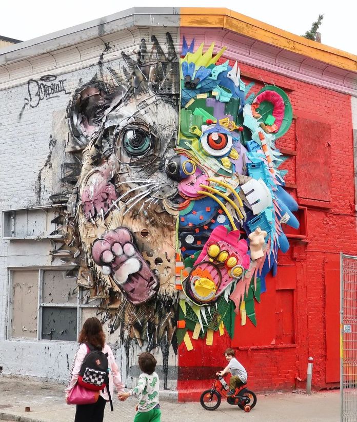 Three-dimensional mural of a hedgehog’s face on a street corner in Montreal, Canada by Bordalo II. The left half is made from dull metal, wires, and industrial waste, while the right half is constructed from colorful plastic trash like toy parts, containers, and helmets. The background wall is painted white on one side and red on the other. A child on a bike and pedestrians walk past the sculpture.