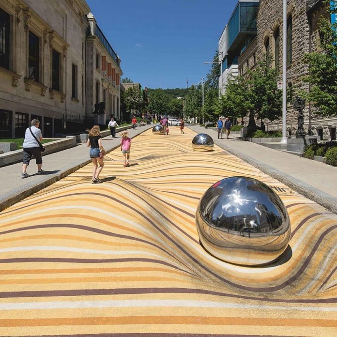 Interactive urban art installation in Montreal featuring a wavy yellow-striped surface with large chrome spheres, surrounded by pedestrians walking and children playing.